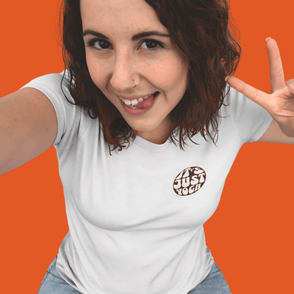 Smiling woman in a white "It's Just Yoga" tee giving a peace sign against an orange background. Perfect for yoga lovers.